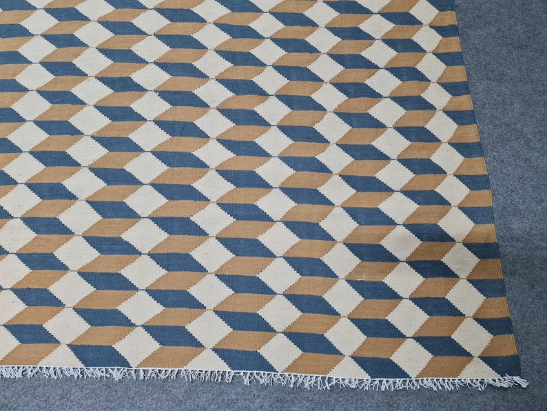 May include: Geometric patterned rug featuring a repeating cube design in blue, tan, and off-white. The rug has a fringed edge and a textured appearance, suitable for home decor.