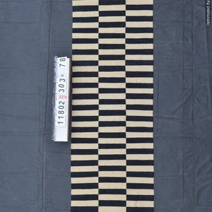 May include: A rectangular rug with a repeating pattern of alternating black and beige horizontal stripes. The rug measures approximately 30 inches by 78 inches. The rug is made of a woven material and is displayed on a dark gray surface.