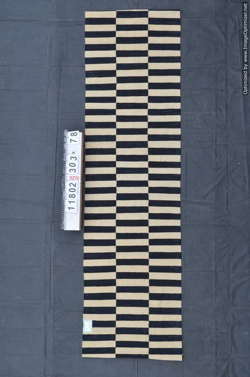 May include: A rectangular rug with a repeating pattern of alternating black and beige horizontal stripes. The rug measures approximately 30 inches by 78 inches. The rug is made of a woven material and is displayed on a dark gray surface.