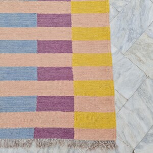 May include: A rectangular rug with a geometric pattern of horizontal stripes in pastel colors. The rug features alternating sections of light pink, blue, purple, and yellow stripes. The rug has a fringed edge.