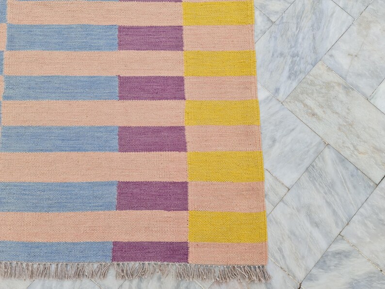 May include: A rectangular rug with a geometric pattern of horizontal stripes in pastel colors. The rug features alternating sections of light pink, blue, purple, and yellow stripes. The rug has a fringed edge.
