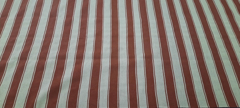 May include: A textile with a repeating pattern of vertical stripes. The stripes alternate between a muted red-brown color and a light beige-white. The fabric appears to be a woven material, possibly cotton or linen.