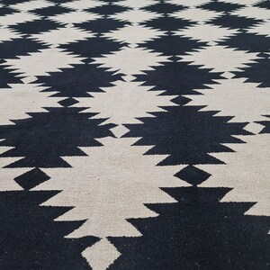 Puede incluir: Una alfombra grande y tejida con un patr&oacute;n geom&eacute;trico repetitivo. El dise&ntilde;o presenta formas de diamante negras y blanquecinas alternas, creando una superficie audaz y texturizada. La construcci&oacute;n de la alfombra sugiere un revestimiento de suelo duradero y decorativo.