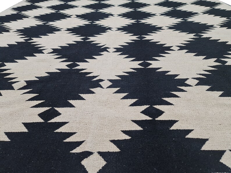 Puede incluir: Una alfombra grande y tejida con un patr&oacute;n geom&eacute;trico repetitivo. El dise&ntilde;o presenta formas de diamante negras y blanquecinas alternas, creando una superficie audaz y texturizada. La construcci&oacute;n de la alfombra sugiere un revestimiento de suelo duradero y decorativo.
