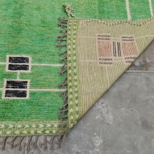 May include: A green and white rectangular rug with a geometric design. The rug features a bright green field with white lines and black rectangular shapes. The edge has a decorative border and tassels. The rug is partially folded over, revealing the underside.