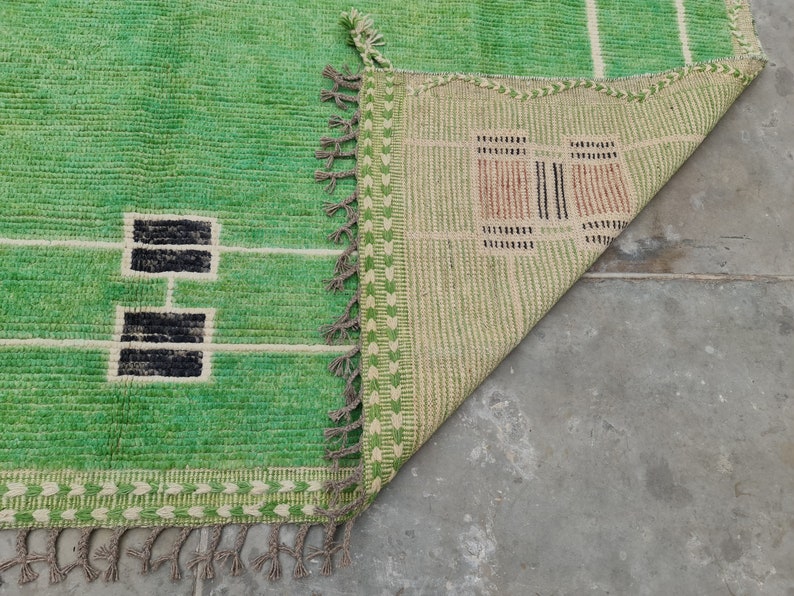 May include: A green and white rectangular rug with a geometric design. The rug features a bright green field with white lines and black rectangular shapes. The edge has a decorative border and tassels. The rug is partially folded over, revealing the underside.
