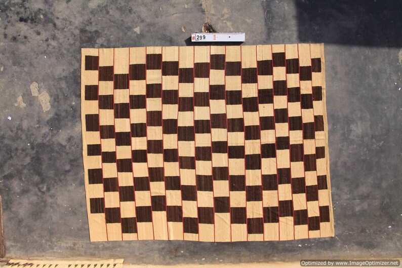 May include: A woven textile with a checkerboard pattern in shades of beige and dark brown. The squares are outlined with thin red lines. The textile is rectangular and appears to be handmade. The tag in the image reads "299".
