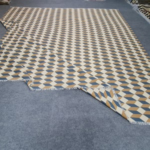 May include: A large, rectangular rug with a repeating geometric pattern of blue, tan, and cream-colored three-dimensional cubes. The rug has a fringed edge and is displayed on a gray surface. The design creates an optical illusion.
