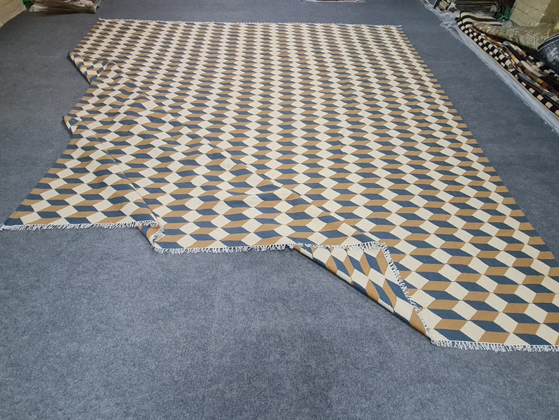 May include: A large, rectangular rug with a repeating geometric pattern of blue, tan, and cream-colored three-dimensional cubes. The rug has a fringed edge and is displayed on a gray surface. The design creates an optical illusion.