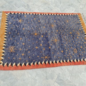 May include: A rectangular rug with a navy blue field scattered with orange starburst designs. The border is a combination of orange, black, and white zigzags, with a tan outer edge and fringe.
