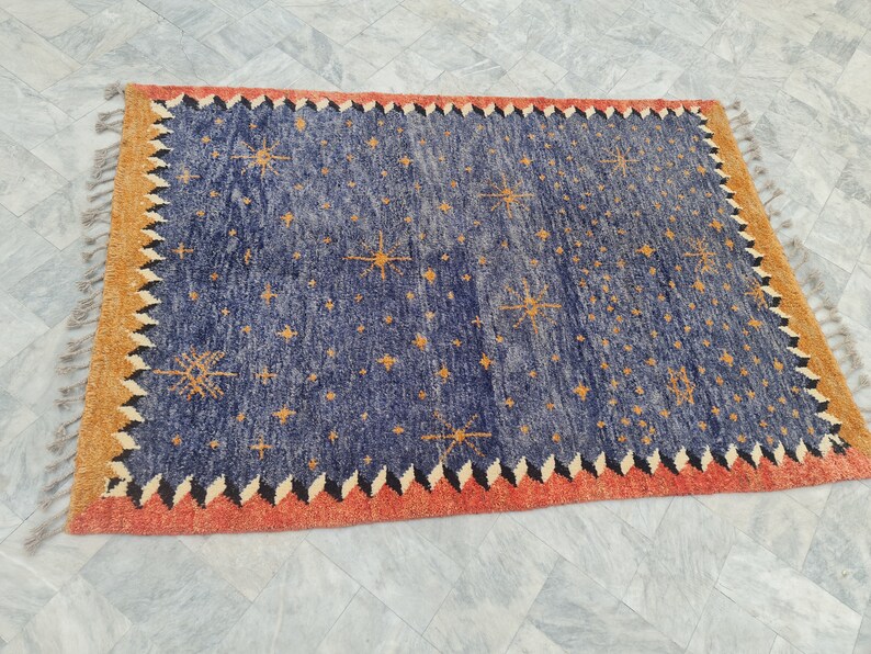 May include: A rectangular rug with a navy blue field scattered with orange starburst designs. The border is a combination of orange, black, and white zigzags, with a tan outer edge and fringe.