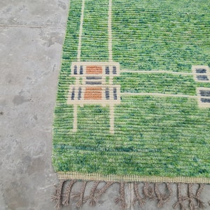 Puede incluir: Una alfombra verde y blanca estampada con una superficie texturizada. La alfombra presenta un dise&ntilde;o con formas geom&eacute;tricas beige, naranja y azul marino. La alfombra tiene una franja a lo largo del borde inferior.
