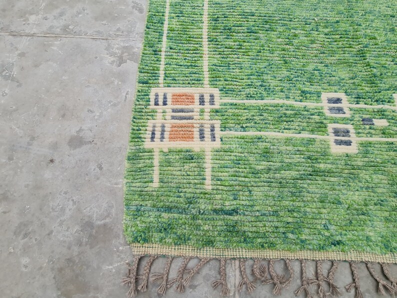 Puede incluir: Una alfombra verde y blanca estampada con una superficie texturizada. La alfombra presenta un dise&ntilde;o con formas geom&eacute;tricas beige, naranja y azul marino. La alfombra tiene una franja a lo largo del borde inferior.