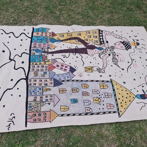 Op de afbeelding: Handgeweven tapijt met een grillig ontwerp met kleurrijke gebouwen, een figuur met een hoed en abstracte elementen. Het tapijt heeft een cr&egrave;mekleurige achtergrond met zwarte en pastelkleurige accenten. Het kunstwerk is linksonder gesigneerd.