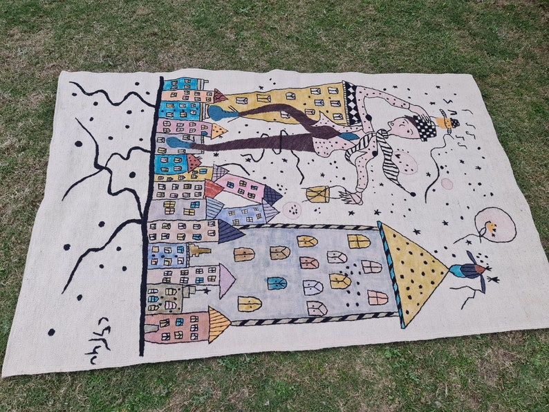Op de afbeelding: Handgeweven tapijt met een grillig ontwerp met kleurrijke gebouwen, een figuur met een hoed en abstracte elementen. Het tapijt heeft een cr&egrave;mekleurige achtergrond met zwarte en pastelkleurige accenten. Het kunstwerk is linksonder gesigneerd.