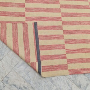 May include: A rectangular rug with a striped pattern in alternating shades of red and beige. The rug is partially rolled up, revealing a dark blue border on the edge. The rug is made of a woven material.