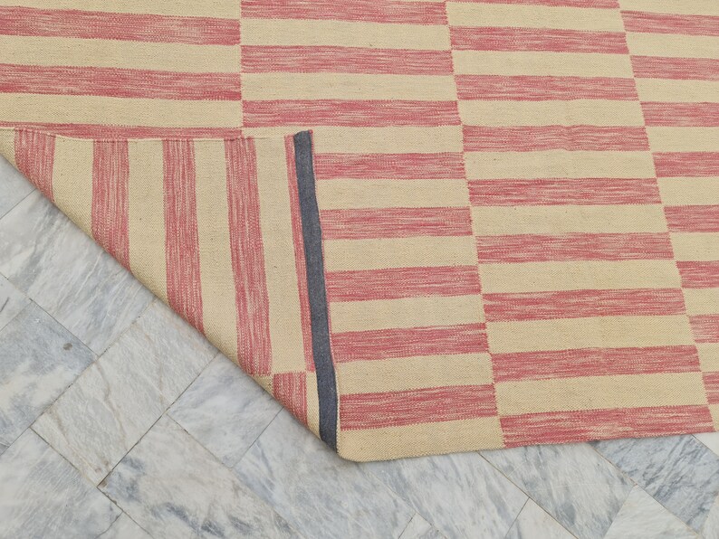 May include: A rectangular rug with a striped pattern in alternating shades of red and beige. The rug is partially rolled up, revealing a dark blue border on the edge. The rug is made of a woven material.