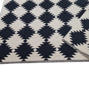 Puede incluir: Una alfombra rectangular con un patr&oacute;n geom&eacute;trico repetido de formas de diamante negras sobre un fondo crema. La alfombra tiene un borde con flecos y est&aacute; parcialmente doblada.