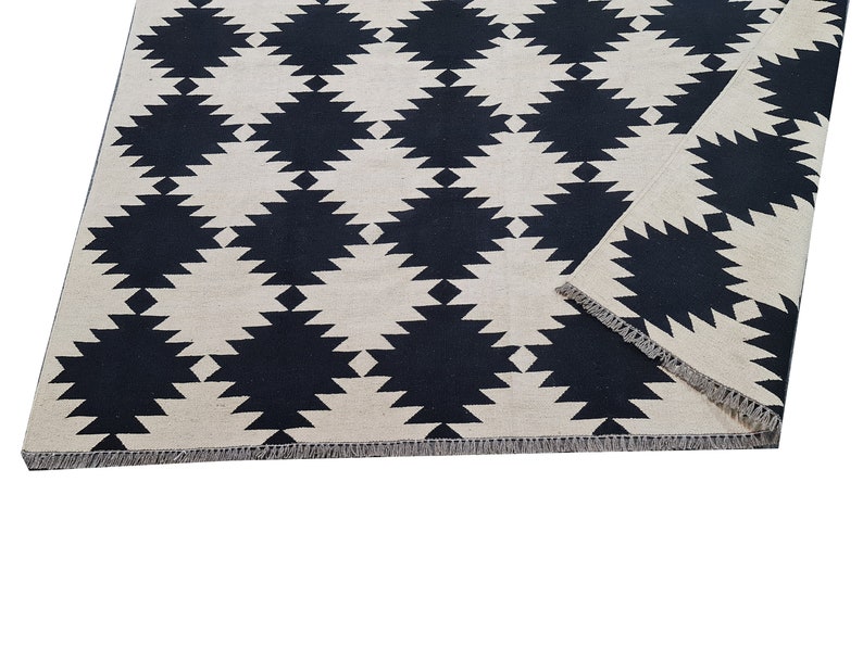 Puede incluir: Una alfombra rectangular con un patr&oacute;n geom&eacute;trico repetido de formas de diamante negras sobre un fondo crema. La alfombra tiene un borde con flecos y est&aacute; parcialmente doblada.