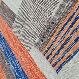 May include: A colorful woven rug with a geometric pattern. The rug features stripes in orange, blue, brown, and white. The rug has a fringed edge and is made of a textured material.