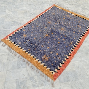 May include: Rectangular blue rug with a starburst pattern and a fringed edge. The rug features a border with orange, black, and white zigzag designs. The rug's dimensions are approximately 5 x 7 feet.