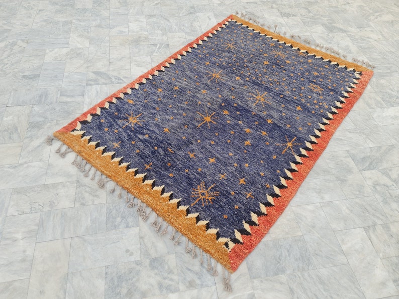 May include: Rectangular blue rug with a starburst pattern and a fringed edge. The rug features a border with orange, black, and white zigzag designs. The rug's dimensions are approximately 5 x 7 feet.