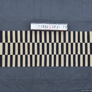 Puede incluir: Una alfombra rectangular con un patrón repetitivo de rayas verticales negras y beige alternas. La alfombra mide aproximadamente 770 cm de largo y 198 cm de ancho. Una etiqueta con el número "11802" es visible. La alfombra se muestra sobre una superficie gris oscuro.