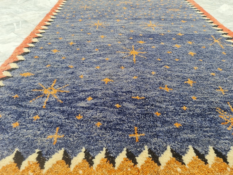 May include: A blue rug with a celestial design. The rug features a pattern of yellow stars scattered across the blue field. The border has a repeating pattern of orange, black, and white shapes.