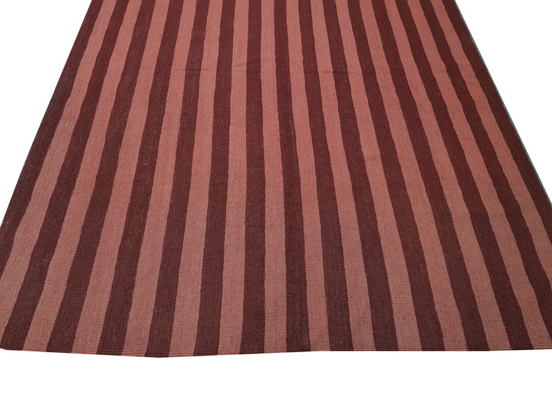 May include: A large, rectangular rug with a striped pattern. The rug features alternating vertical stripes of dark and light reddish-brown colors. The rug is made of a woven material and is displayed on a white background.