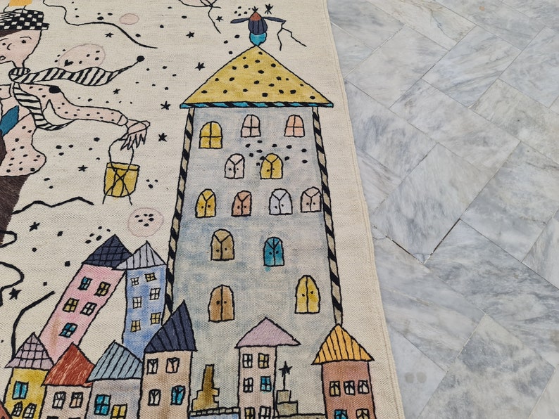 Op de afbeelding: Een handgetekend tapijt met een grillig stadslandschapontwerp. Het heeft kleurrijke gebouwen, een hoge toren en een figuur met een sjaal. Een vogel zit op het dak van de toren. Het tapijt heeft een cr&egrave;mekleurige achtergrond.