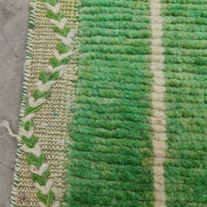 May include: Close-up of a green and white striped rug. The rug has a textured, woven appearance with a central white stripe and green stripes. A decorative border with green and white leaf-like patterns is visible on the left side.