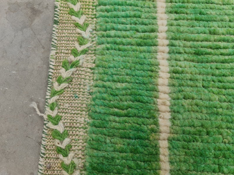 May include: Close-up of a green and white striped rug. The rug has a textured, woven appearance with a central white stripe and green stripes. A decorative border with green and white leaf-like patterns is visible on the left side.