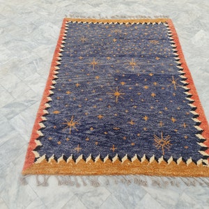 May include: Rectangular blue rug with a star pattern and a red and orange border. The rug has a fringed edge and is likely made of wool or a similar material. The design features multiple starbursts.