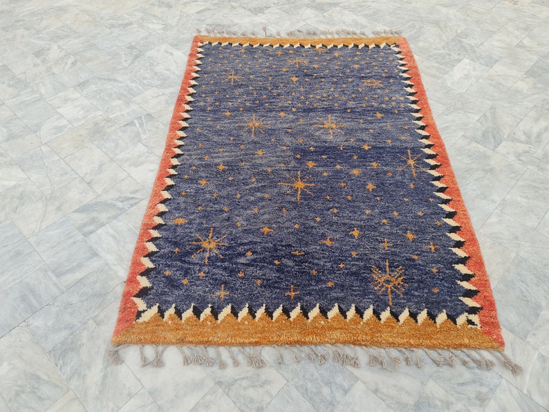 May include: Rectangular blue rug with a star pattern and a red and orange border. The rug has a fringed edge and is likely made of wool or a similar material. The design features multiple starbursts.
