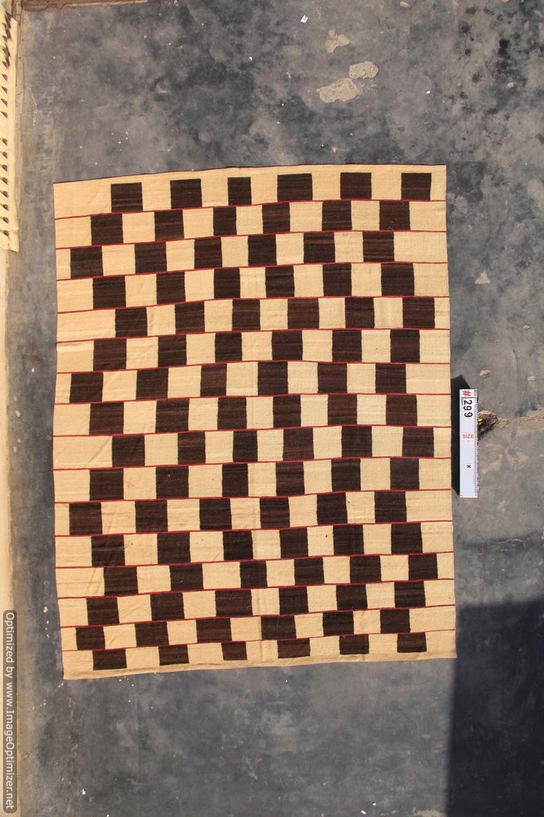 May include: A rectangular woven rug with a repeating pattern of alternating dark brown and beige squares. Thin red lines separate the horizontal rows. The rug is displayed on a gray surface.