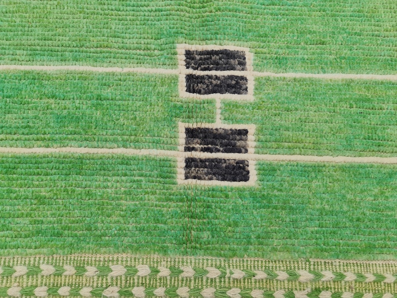 May include: Close-up of a green rug with a textured surface. The rug features a design with black rectangular shapes connected by white lines. A decorative border with a repeating pattern of green and white shapes is at the bottom.