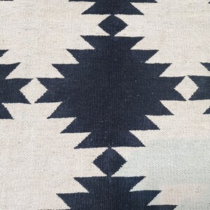 Puede incluir: Primer plano de una alfombra tejida con un patr&oacute;n geom&eacute;trico repetitivo. El dise&ntilde;o presenta formas de diamante azul marino oscuro y blanco roto alternadas. La alfombra tiene una superficie texturizada.