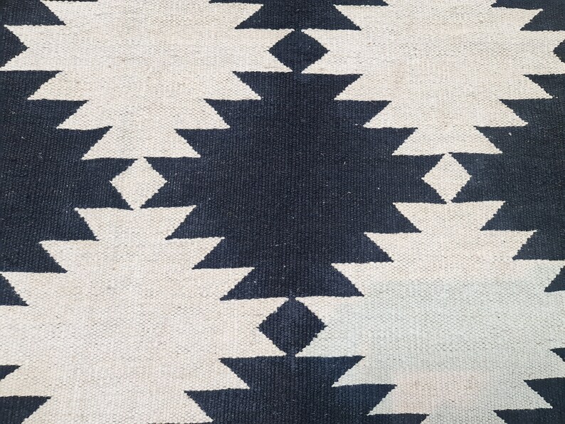 Puede incluir: Primer plano de una alfombra tejida con un patr&oacute;n geom&eacute;trico repetitivo. El dise&ntilde;o presenta formas de diamante azul marino oscuro y blanco roto alternadas. La alfombra tiene una superficie texturizada.