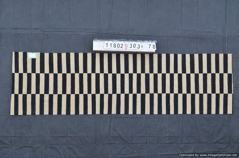 May include: A rectangular rug with a repeating pattern of alternating black and beige vertical stripes. The rug is approximately 303 inches long and 78 inches wide. A tag with numbers is placed on top of the rug.