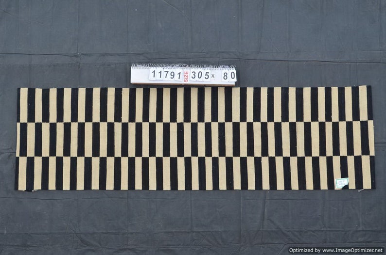 May include: A rectangular rug with a repeating pattern of black and beige vertical stripes. The rug measures approximately 120 inches long and 31.5 inches wide. The rug is made of a woven material and is displayed on a dark gray surface.