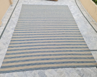 May include: A large rectangular rug with horizontal stripes in alternating shades of blue and beige. The rug is made of a woven material and is placed on a light-colored tiled surface. The rug's design is simple and modern.