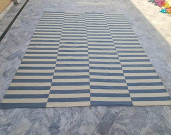 Puede incluir: Una alfombra rectangular grande con un patrón geométrico de rayas horizontales azules y crema alternas. La alfombra está hecha de un material tejido y está colocada sobre una superficie de baldosas grises.
