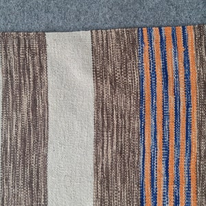 May include: A woven rug with vertical stripes in beige, brown, orange, and blue. The rug has a fringed edge and a textured appearance. The color scheme is neutral with a pop of color from the stripes.