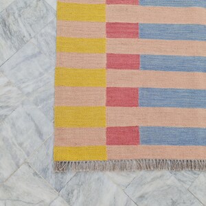 May include: A colorful woven rug with a geometric pattern. The rug features horizontal stripes in yellow, pink, red, and blue. The rug has a fringed edge and is displayed on a marble tile surface.
