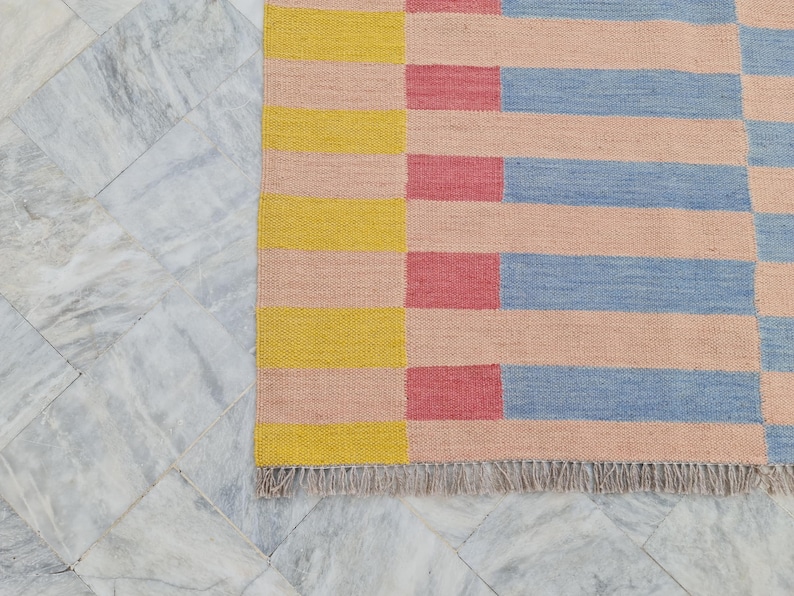 May include: A colorful woven rug with a geometric pattern. The rug features horizontal stripes in yellow, pink, red, and blue. The rug has a fringed edge and is displayed on a marble tile surface.