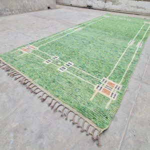 Puede incluir: Una alfombra rectangular verde con un dise&ntilde;o geom&eacute;trico. La alfombra presenta un campo verde claro con un borde blanco y cuadrados decorativos en beige, azul marino y naranja. La alfombra tiene flecos en un extremo.