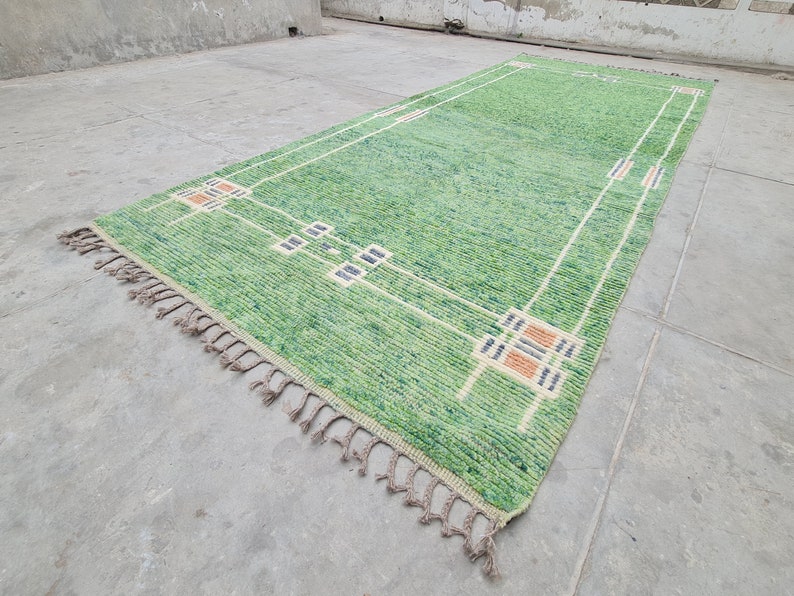 Puede incluir: Una alfombra rectangular verde con un dise&ntilde;o geom&eacute;trico. La alfombra presenta un campo verde claro con un borde blanco y cuadrados decorativos en beige, azul marino y naranja. La alfombra tiene flecos en un extremo.