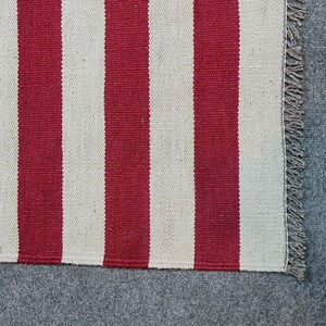 Puede incluir: Primer plano de una alfombra a rayas rojas y blancas. La alfombra tiene gruesas rayas verticales rojas y blancas. El borde de la alfombra tiene un detalle de flecos. La alfombra est&aacute; hecha de un material tejido.