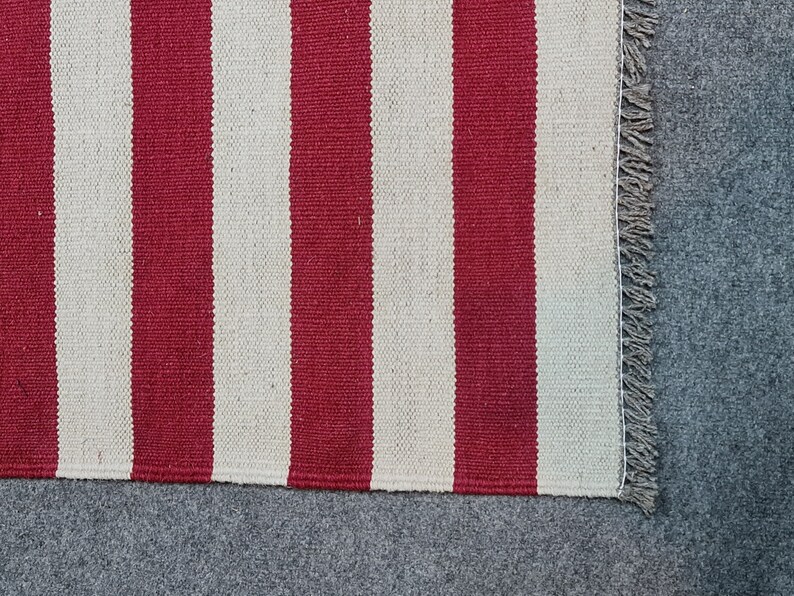 Puede incluir: Primer plano de una alfombra a rayas rojas y blancas. La alfombra tiene gruesas rayas verticales rojas y blancas. El borde de la alfombra tiene un detalle de flecos. La alfombra est&aacute; hecha de un material tejido.