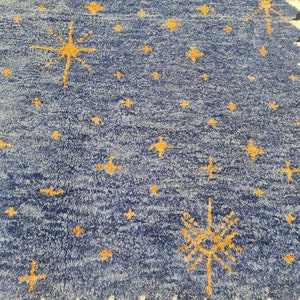 May include: A blue rug with a pattern of yellow stars and starbursts. The rug has a textured surface and a border with orange, black, and white geometric designs. The rug is likely used for home decor.